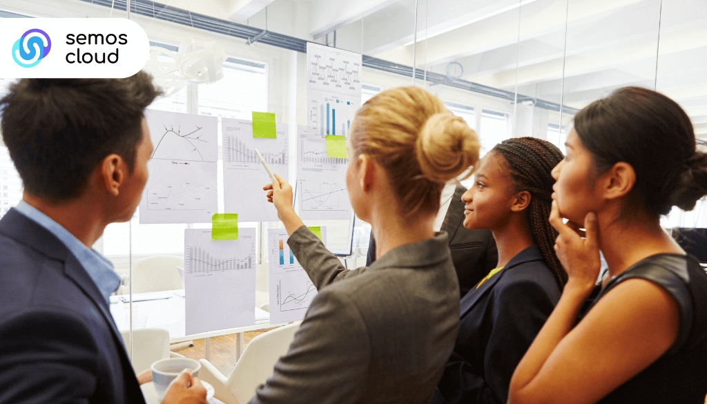 Alt text: Four professionals in an office review printed charts and graphs taped to a glass wall while one points with a pen; the Semos Cloud logo is in the top-left corner.